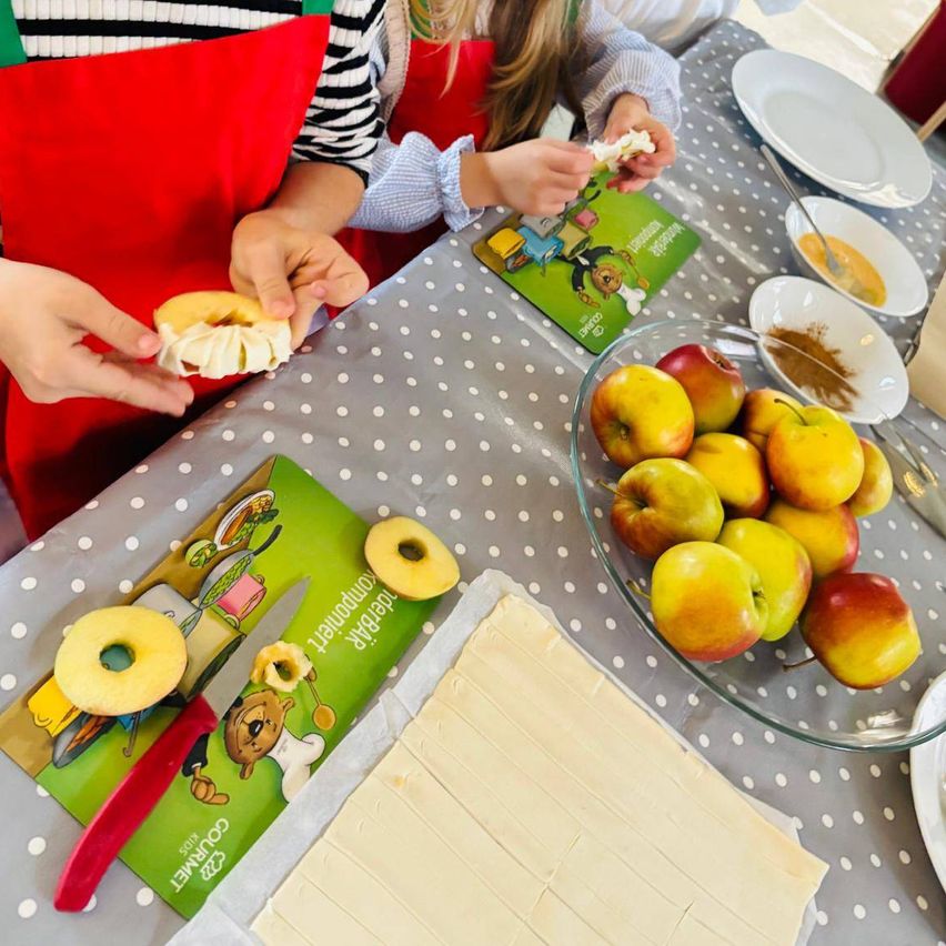 Two children in aprons are making apple pastries. One holds a cut apple and a dough ring, the other has a pastry cutter. A table with apples, ingredients, and a book is covered with a polka-dot tablecloth.