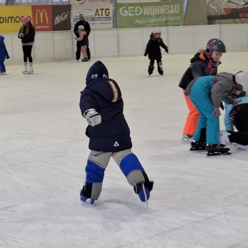 Eine Gruppe von Menschen skat auf einer überdachten Eisbahn. Ein Kind hält einen Hockeyschläger. Andere tragen Helme und Schlittschuhe. Werbung ist an der Wand angebracht.