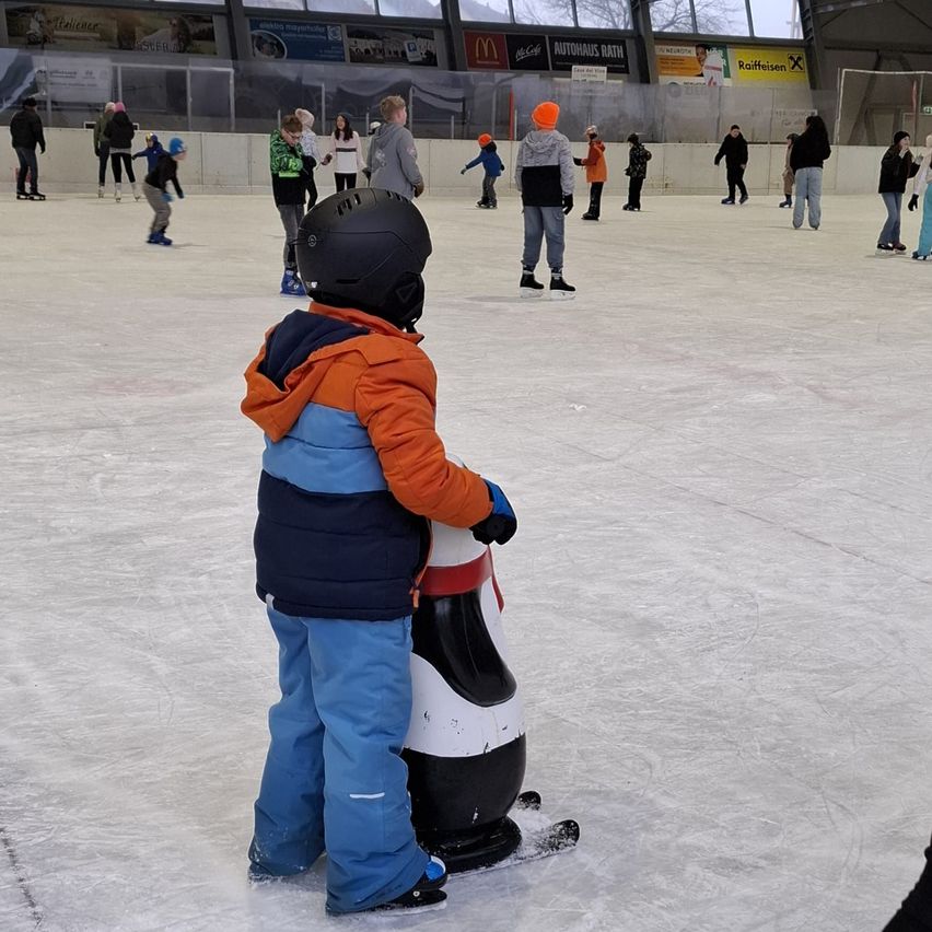 Auf einer Eisbahn steht ein Kind neben einer Pinguin-Skulptur, bekleidet mit blauer Hose und orangefarbener Jacke. Im Hintergrund skaten mehrere Personen.