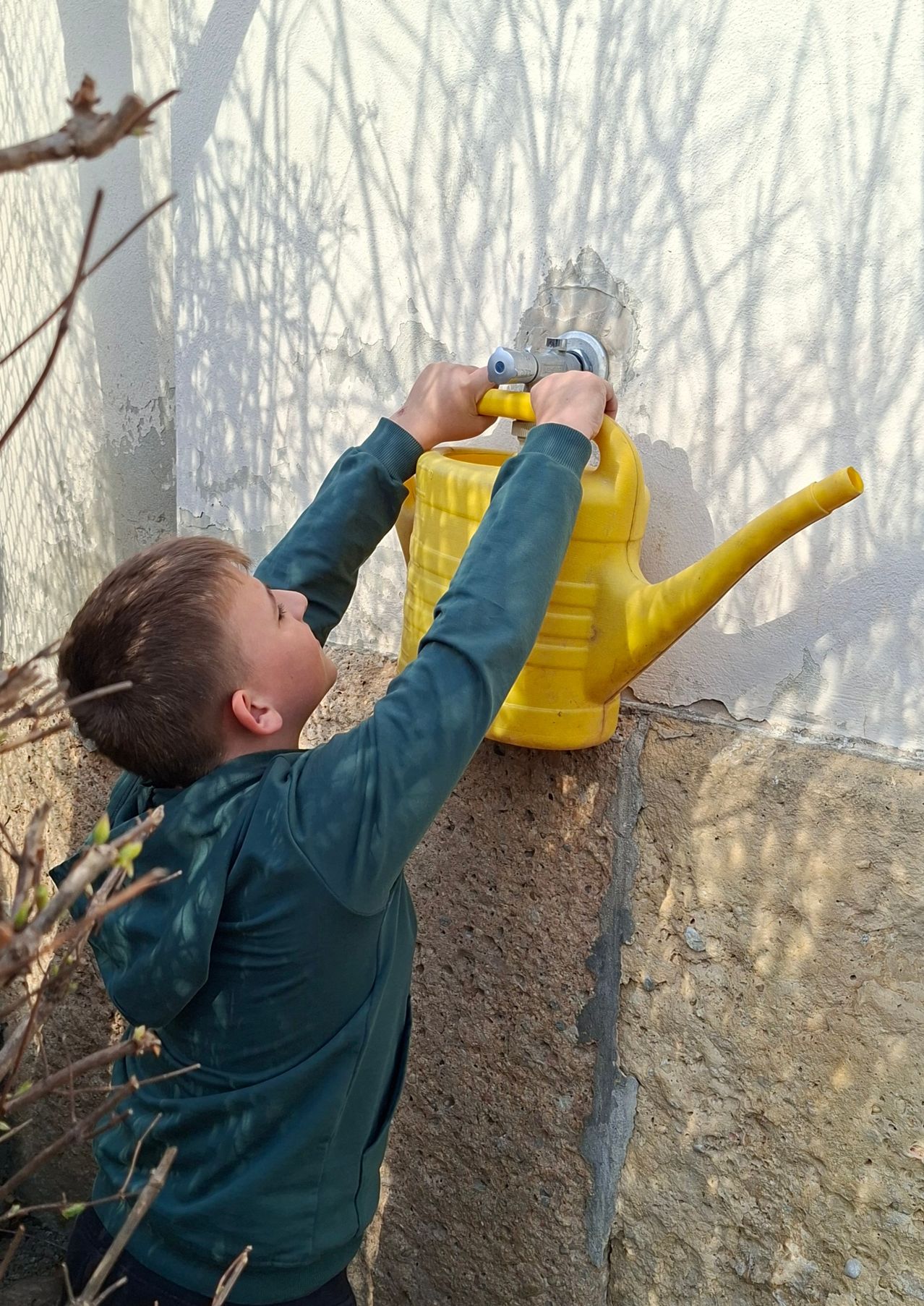 A young boy fills a yellow watering can from a wall-mounted faucet, leaning against a stone wall with shadows cast by trees.