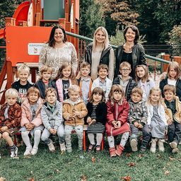 Mehrere junge Kinder posieren für ein Gruppenfoto in einem Park mit Lehrern vor einem Spielplatz.