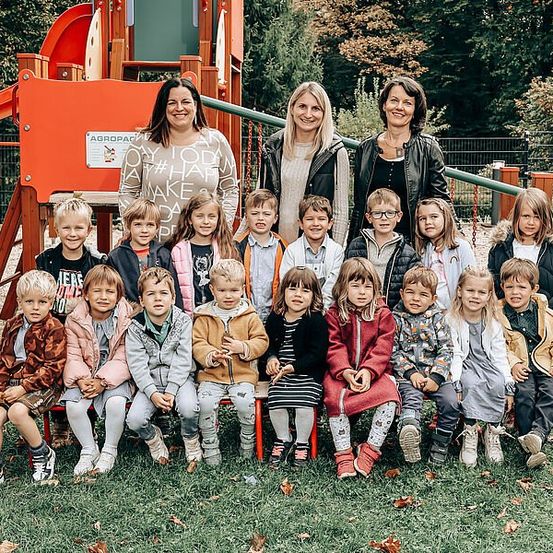 Mehrere junge Kinder posieren für ein Gruppenfoto in einem Park mit Lehrern vor einem Spielplatz.