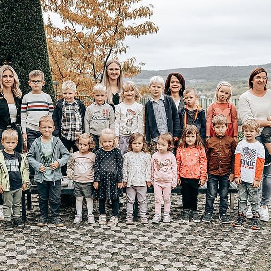 Eine Gruppe von Kindern und Erwachsenen steht in einer Reihe, lächelt und posiert für ein Foto. Sie befinden sich auf einem Kopfsteinpflasterweg, mit einem Baum und einem Zaun hinter ihnen.