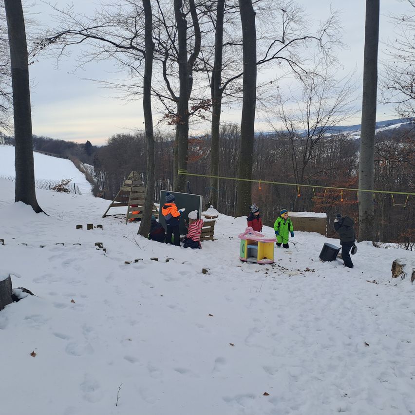 Kinder spielen im Schnee an einem Hang. Einige sitzen auf einer Holzkonstruktion, während andere stehen. Ein Kind trägt Mütze und Handschuhe. Ein Tafel ist gegen einen Baum gelehnt.