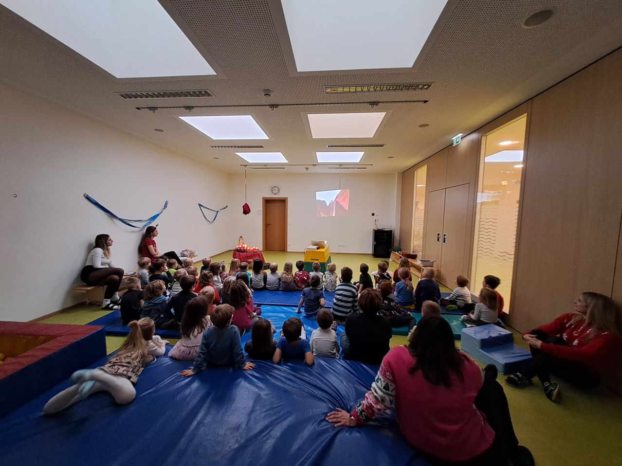 Eine Gruppe von Kindern und Erwachsenen sitzt auf blauen Matten auf dem Boden und nimmt an einer Versammlung teil. Der Raum hat weiße Wände und quadratische Deckenleuchten. An der Wand ist eine Projektionsleinwand angebracht.
