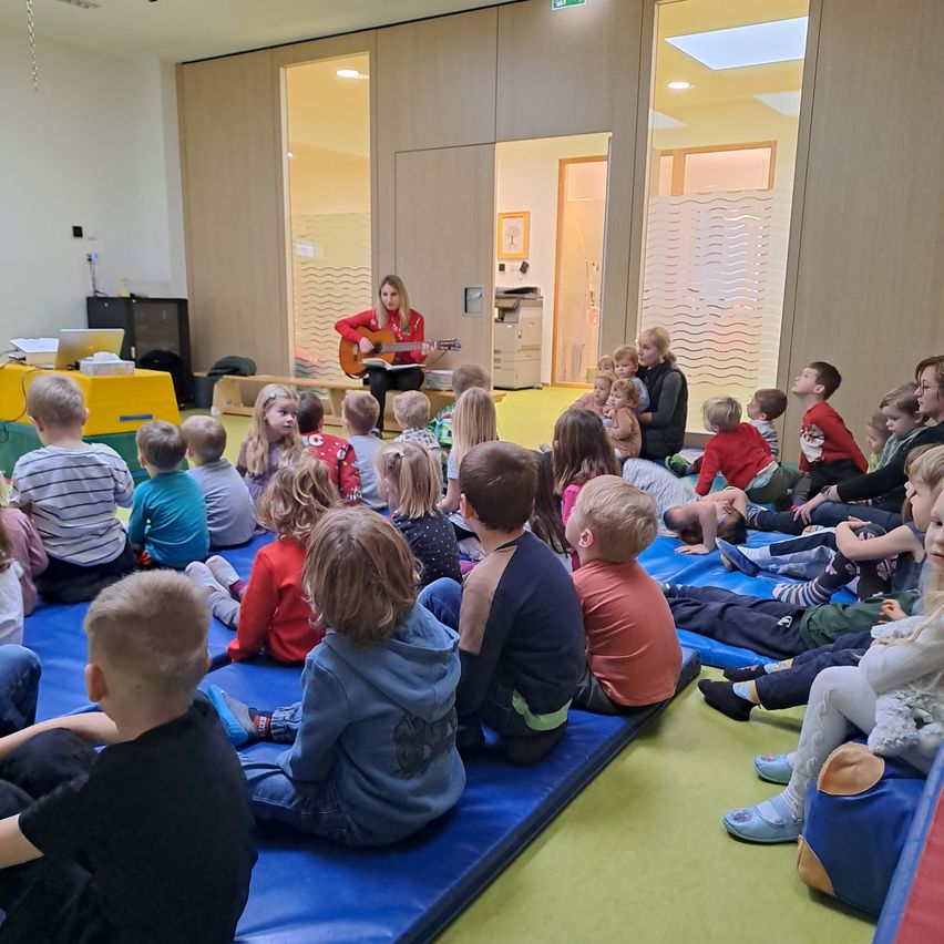 Eine Frau spielt Gitarre vor einer Gruppe von Kindern in einem Raum. Die Kinder sitzen auf blauen Matten auf dem Boden.