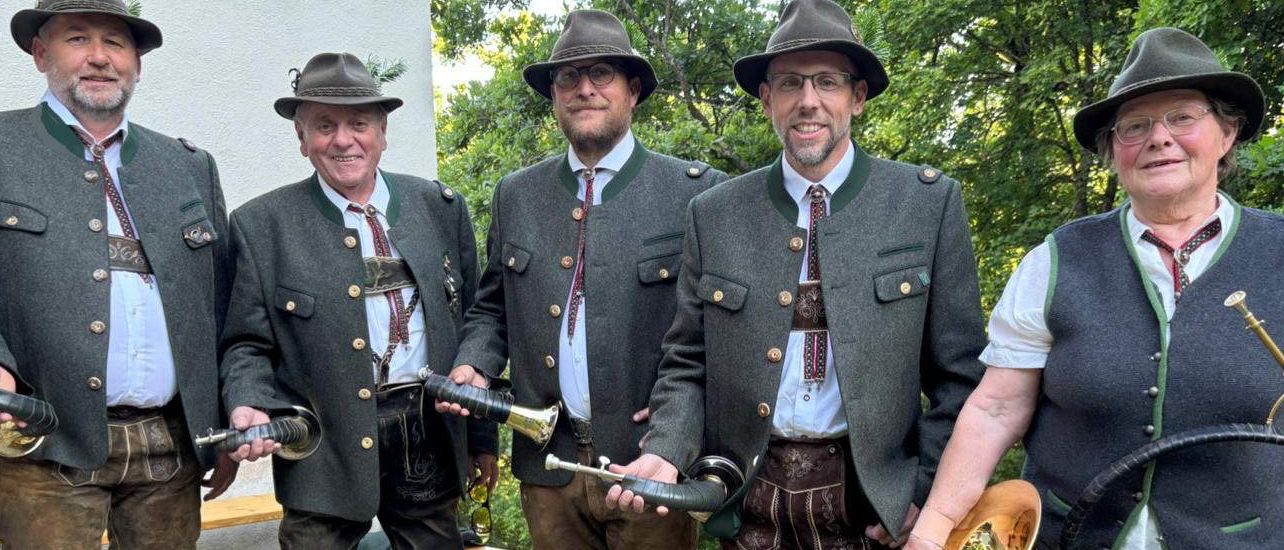 Fünf Männer in traditioneller bayerischer Tracht stehen in einem Grasbereich und halten Messinginstrumente. Hinter ihnen ist eine teilweise sichtbare Holzbank, und Bäume bilden einen üppigen Hintergrund.