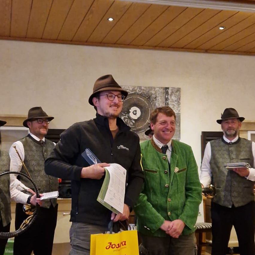 A man in a hat and glasses holds a paper and a yellow bag. Beside him, a smiling man in a green jacket. Behind them, three men in hats, two holding papers. A trumpet is visible in the background.