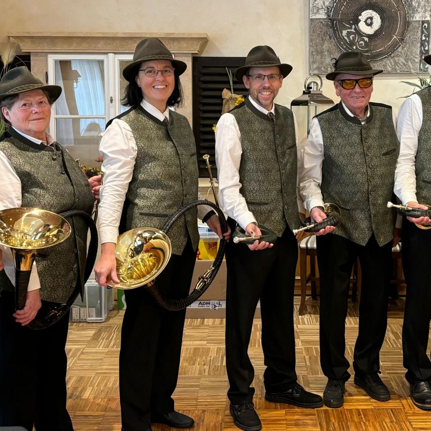 Five people in matching green vests and black pants stand together, smiling and posing for a photo. They all wear hats and hold brass instruments. Behind them is a wall with a framed picture and a window with curtains.