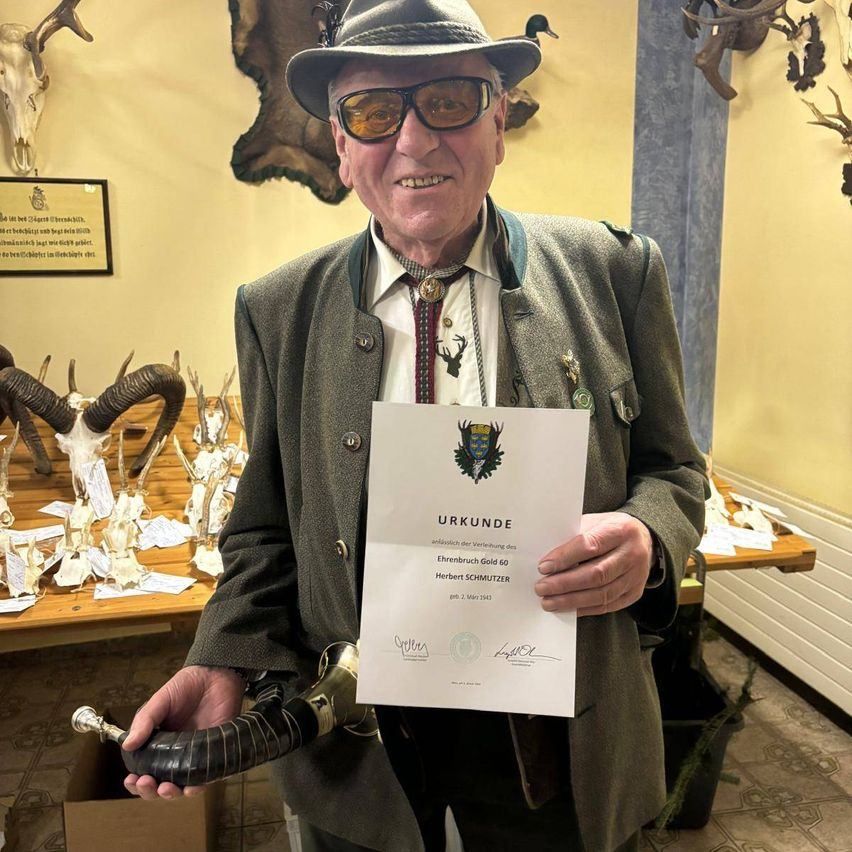 Ein älterer Mann steht und hält ein Zertifikat und einen Horn, lächelt. Hinter ihm steht ein Tisch mit Hirschköpfen.