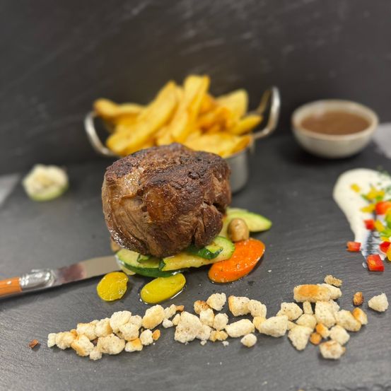Ein gut zubereitetes Steak mit Gemüse und Croutons auf einem Schieferteller, begleitet von einer Schüssel Sauce und einer Frittenschale.