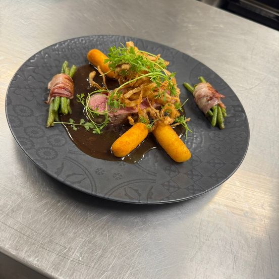 Ein Gourmetgericht mit einem Stück Fleisch, grünen Bohnen und Karotten, garniert mit gebratenen Zwiebeln und Kräutern, serviert auf einem schwarzen Teller mit dunkler Sauce.