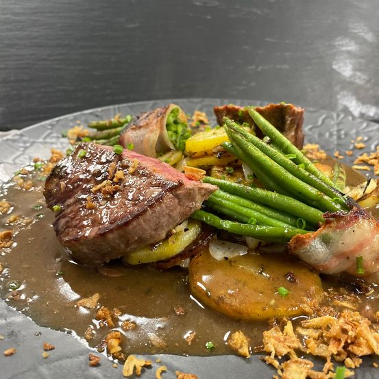 Ein Gourmetgericht mit Steak, grünen Bohnen und Kartoffeln, garniert mit knusprigen Speckstückchen und einer reichhaltigen Sauce, serviert auf einem grauen Teller.