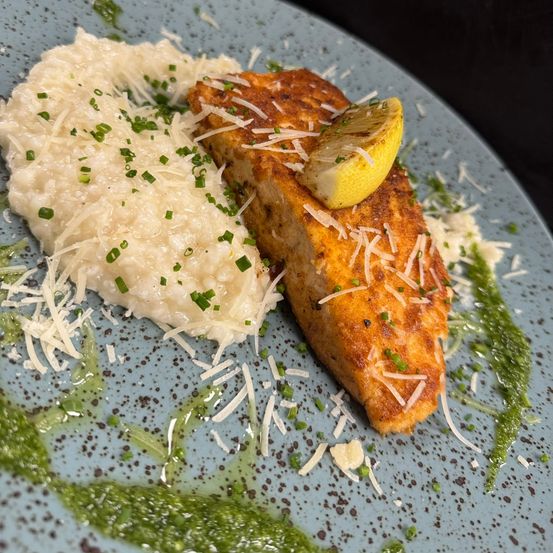 Ein Teller mit einem Stück Lachs, überzogen mit einer Zitronenscheibe und geriebenem Käse, begleitet von cremigem Risotto und grüner Garnitur.