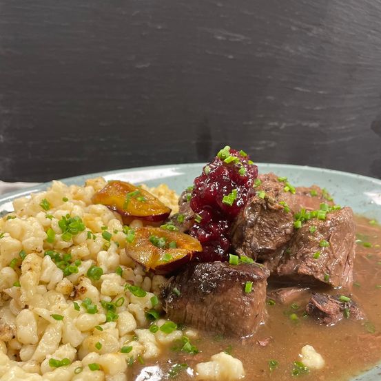 Ein Teller mit Rindergulasch mit Kartoffelpüree, Preiselbeeren und Frühlingszwiebeln. Das Gericht wird auf einem weißen Teller serviert.