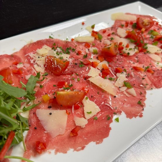 Ein Teller mit dünn geschnittenem Rindfleisch-Carpaccio mit Tomaten, Käse und Kräutern. Das Gericht ist mit Rucola garniert.