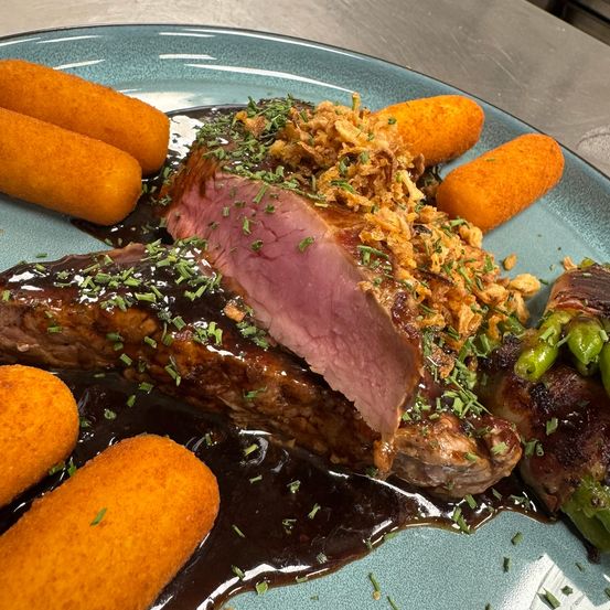 Ein blauer Teller mit geschnittenem Steak, garniert mit Kräutern und knusprigen Zwiebeln, begleitet von einer Beilage aus Spargel und goldenen Kartoffelsticks.