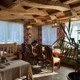 Ein gemütliches Restaurant mit Holzbalken, einem langen Tisch und Stühlen. Der Raum hat weiße Vorhänge und eine Topfpflanze rechts.