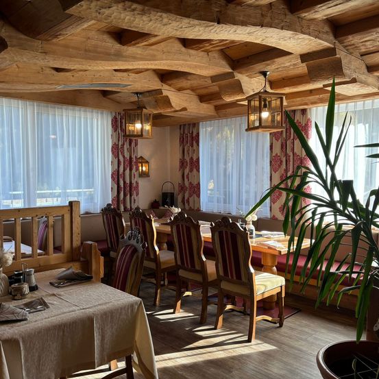 Ein gemütliches Restaurant mit Holzbalken, einem langen Tisch und Stühlen. Der Raum hat weiße Vorhänge und eine Topfpflanze rechts.