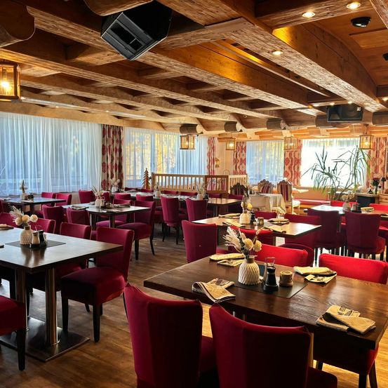 Ein Restaurant mit Holzbalken, roten Stühlen und Tischen. Der Raum ist gut beleuchtet mit Lampen und hat ein großes Fenster mit Vorhängen.