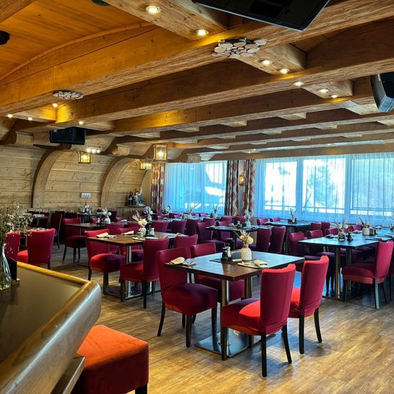 Ein Restaurant mit Holzböden, Holzbalken und roten Stühlen. Die Tische sind mit Blumen und Servietten gedeckt. Der Raum hat große Fenster mit Vorhängen.