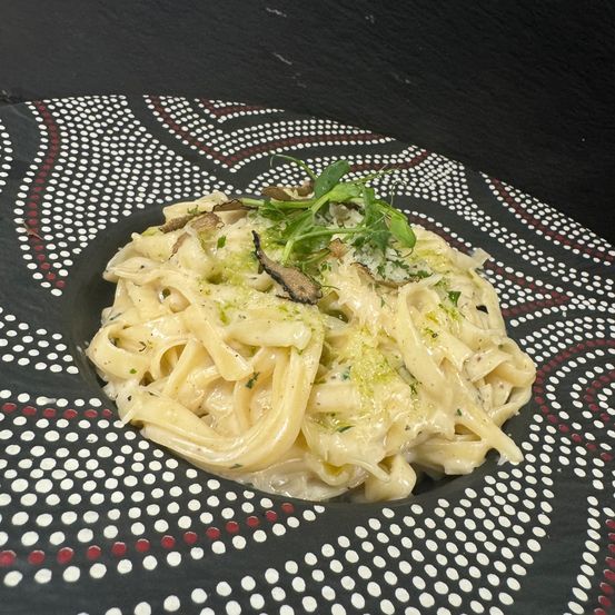 Ein Teller mit cremiger Pasta, grünen Kräutern und Trüffelscheiben obenauf. Die Pasta ist mit einem grünen Kraut und einer Trüffelscheibe garniert.