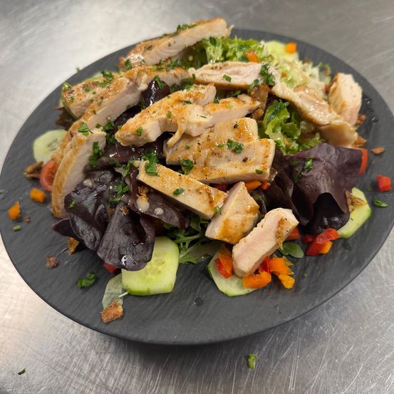 Ein Teller mit gegrilltem Hähnchensalat mit gemischten Salaten, Gurken und roten Paprikaschoten auf einem schwarzen Teller.