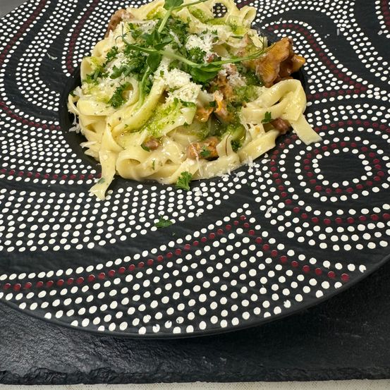 Ein Teller mit Nudeln in grüner Sauce, garniert mit Kräutern und Käse, auf einem gemusterten schwarzen Teller.