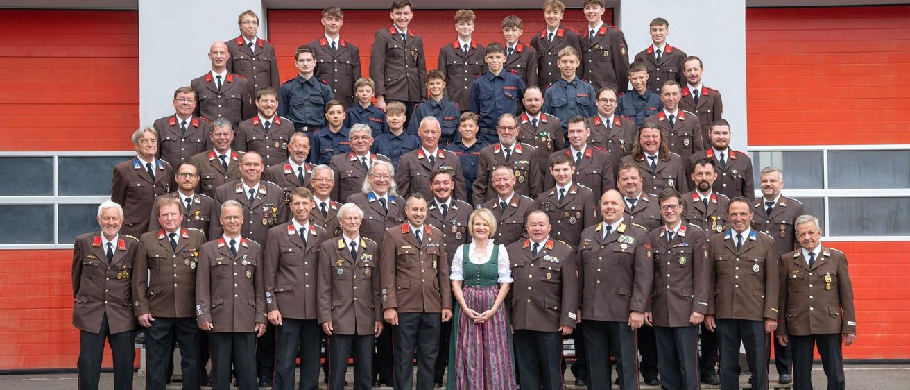 Eine Gruppe von Menschen in Uniformen, einige stehend, einige sitzend, posiert für ein Foto vor einer Wand mit dem Text 'LLIGE FEUERWEHR OBE'.