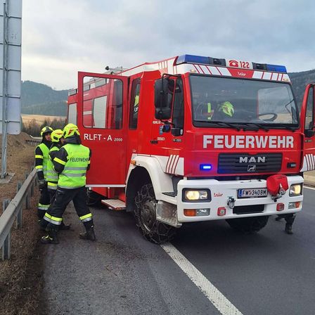 Ein roter Feuerwehrwagen mit der Nummer 122 steht am Straßenrand. Vier Feuerwehrleute stehen neben dem Fahrzeug, einer sitzt im Inneren.