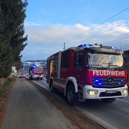 Ein roter Feuerwehrwagen fährt mit einem weiteren Feuerwehrwagen und einem Auto dahinter die Straße entlang. Der Feuerwehrwagen hat blaue Lichter oben und 'Feuerwehr' steht vorne drauf.
