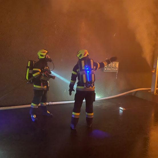Zwei Feuerwehrleute in voller Ausrüstung stehen in einer rauchigen Umgebung. Einer zeigt mit einer Taschenlampe nach vorne, während der andere ein Gerät hält. Sie befinden sich in der Nähe eines Schildes, das 30 Meter und 155 Meter anzeigt.
