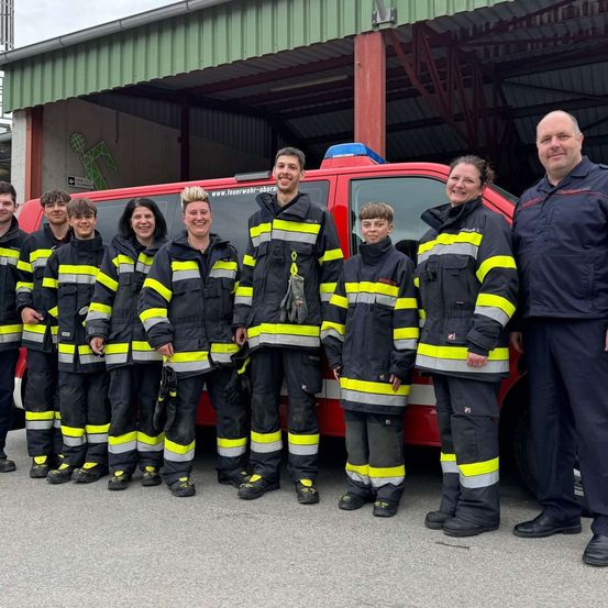 Eine Gruppe von Feuerwehrleuten, einschließlich Kindern, steht vor einem roten Feuerwehrauto und lächelt für ein Foto. Der Hintergrund zeigt ein Feuerwehrhaus mit einem grünen Gebäude.