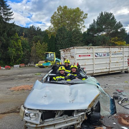 Drei Feuerwehrleute in gelben Helmen posieren in einem demolierten Auto. Dahinter sind ein großer weißer Container mit UWS-Kennzeichnung und ein gelber Walze zu sehen. Bäume und ein Berg im Hintergrund.