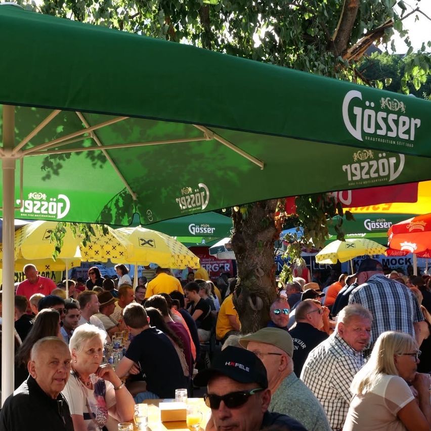 Eine Menschenmenge versammelt sich in einem Außenbereich unter grünen und gelben Schirmen. Die Schirme haben Logos. Ein Baumstamm ist sichtbar.