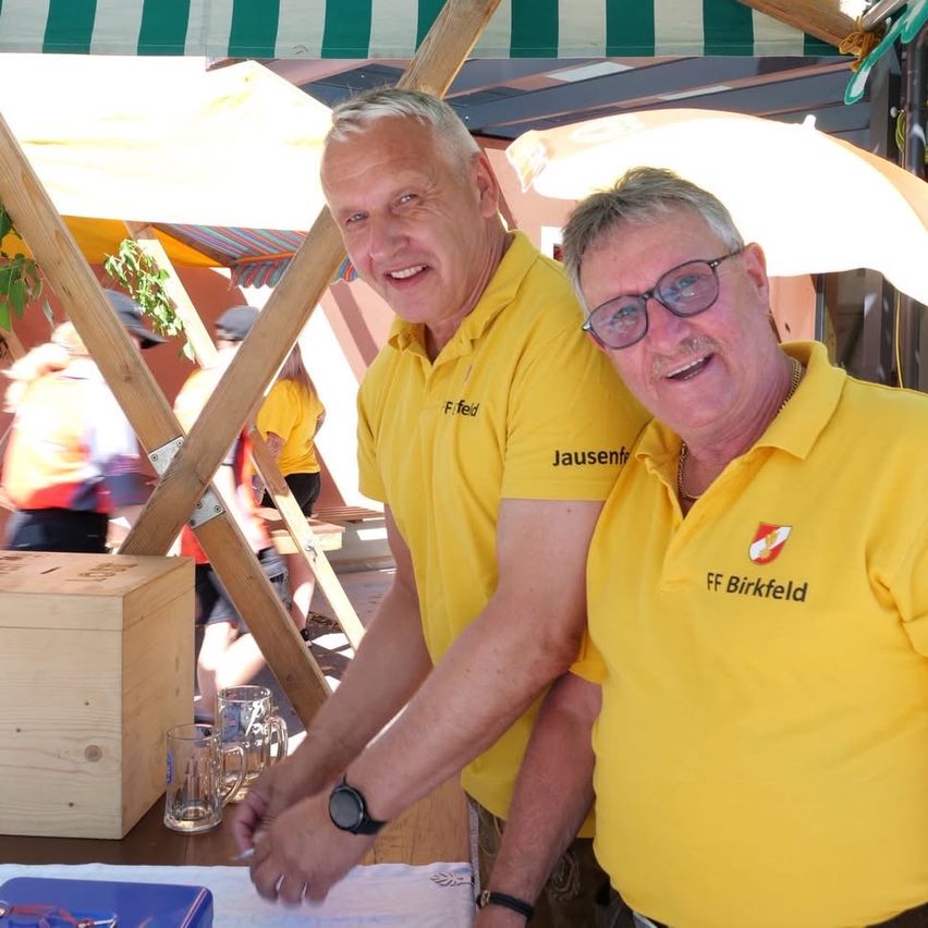 Zwei Männer in gelben Poloshirts stehen lächelnd, einer hält einen Stift. Hinter ihnen steht eine Holzkiste und Gläser auf einem Tisch, und Menschen gehen im Hintergrund vorbei.