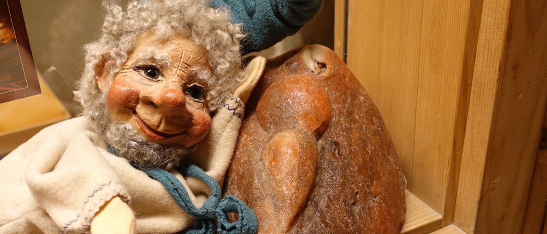 Eine Puppe mit weißem Bart hält ein großes Stück Brot mit einem Biss darauf. Die Puppe hat ein blaues Hemd und eine weiße Schürze.