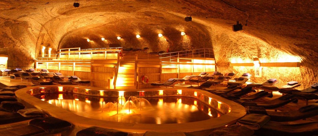 Ein runder Pool mit Liegestühlen und Bänken ist in einer höhlenartigen Umgebung von warmen Lichtern beleuchtet. Eine Fontäne befindet sich in der Mitte, und eine Treppe führt zu einer Holzplattform.