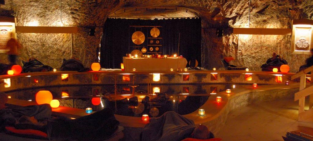 Eine schwach beleuchtete Höhle mit einem Pool und einem Loungebereich, geschmückt mit Kerzen und einer Bühne mit hängenden Gongs.