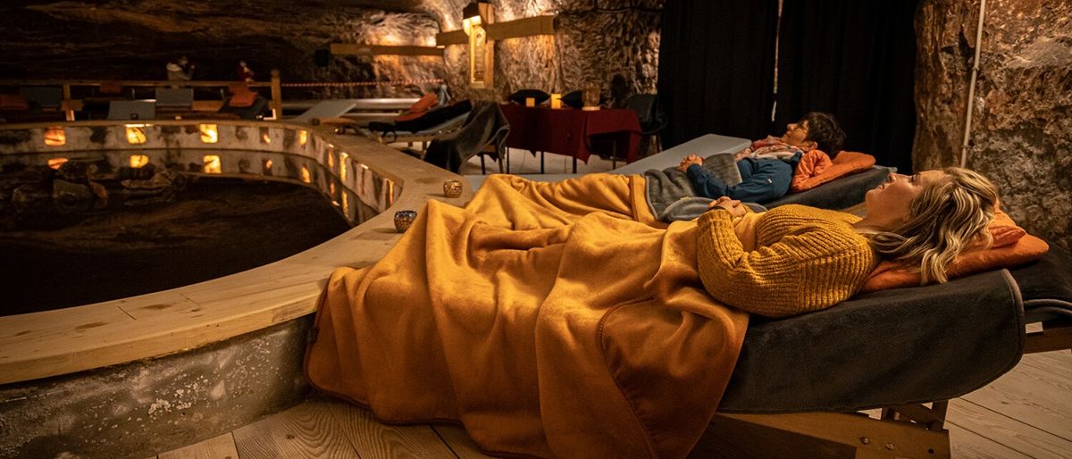 Zwei Personen liegen auf einem Bett, das mit einer Decke bedeckt ist, in einem schwach beleuchteten, höhlenartigen Raum. Mehrere Liegestühle und ein Tisch mit roter Tischdecke sind im Raum aufgestellt.