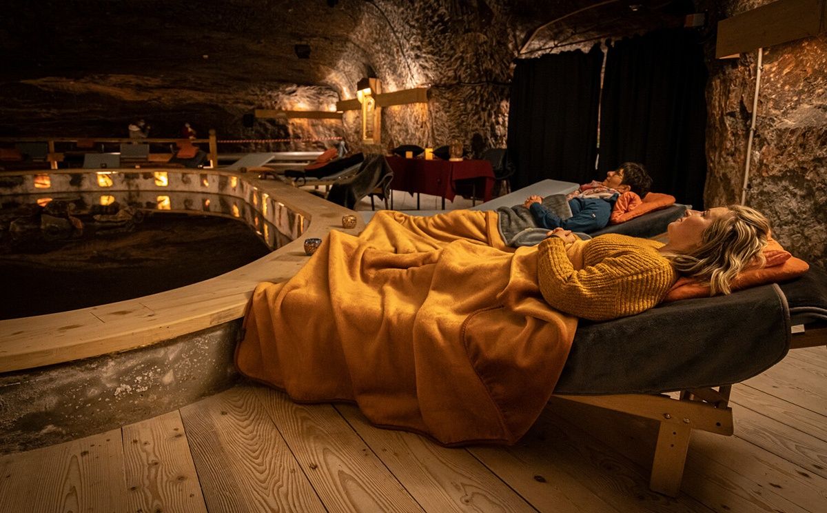 Zwei Personen liegen auf einem Bett in einem dunkel beleuchteten, unterirdischen Raum mit Steinwänden. Eine Person trägt einen gelben Pullover. Ein Tisch und Stühle sind in der Nähe.