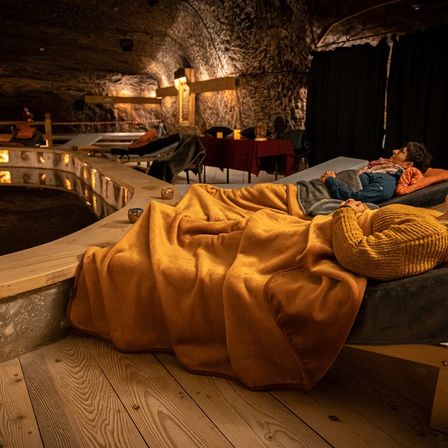 Zwei Personen liegen auf einem Bett in einem dunkel beleuchteten, unterirdischen Raum mit Steinwänden. Eine Person trägt einen gelben Pullover. Ein Tisch und Stühle sind in der Nähe.