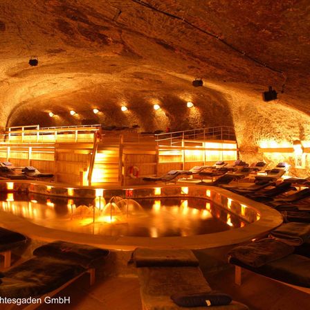 Ein unterirdisches Spa mit einem großen runden Pool, der von warmen Lichtern beleuchtet wird. Holzliegen umgeben den Pool mit einem Brunnen in der Mitte.