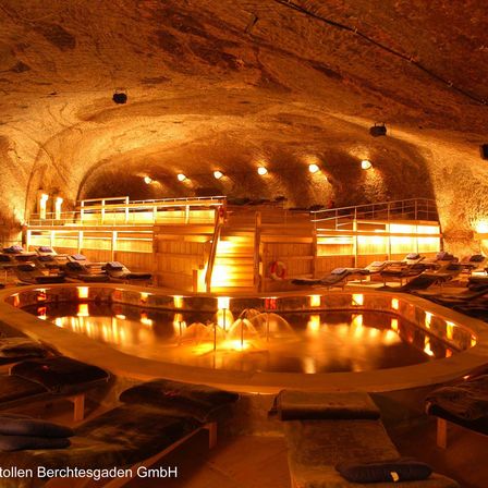 Ein kreisförmiger Spa-Pool, der von warmen Lichtern beleuchtet wird, umgeben von Liegestühlen und einer Holztreppe. Die Decke besteht aus Stein mit hängenden Lampen.