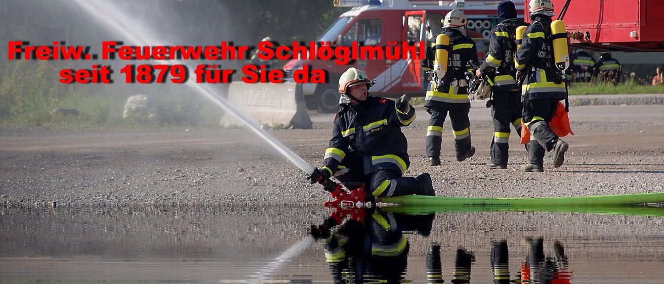 Feuerwehrleute sind bei einer Trainingsübung. Ein Feuerwehrmann sprüht Wasser, während ein anderer daneben steht. Der Text im Bild lautet Wehrwehr Schloglmuhl.