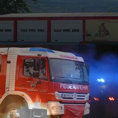 Ein Feuerwehrauto, gekennzeichnet mit 'Feuerwehr', steht vor einer Feuerwehrstation mit drei Garagen. Der LKW ist rot und weiß, und hinten gibt es blaue Beleuchtung.