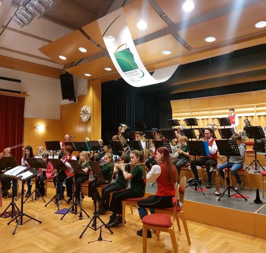 Eine Gruppe junger Musiker in grünen Uniformen spielt verschiedene Instrumente, darunter Trompeten, Posaunen und Flöten, auf einer Bühne mit Notenständern und einem Hintergrund mit Logo.