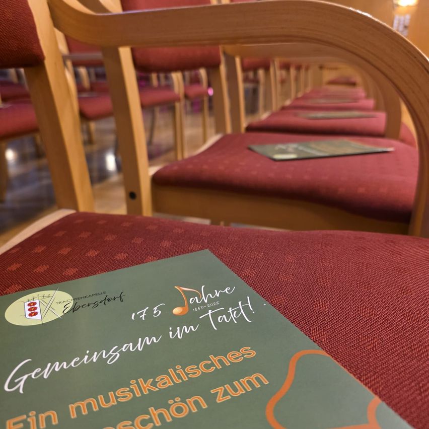 Eine Nahaufnahme einer Broschüre mit dem Titel 'Gemeinsam im Takt' liegt auf einem rot gepolsterten Stuhl. Dahinter ist eine Reihe von Holzstühlen mit roten Bezügen zu sehen. Die Stühle sind in einer Reihe angeordnet. Ein Buch liegt auf dem Sitz eines der Stühle.