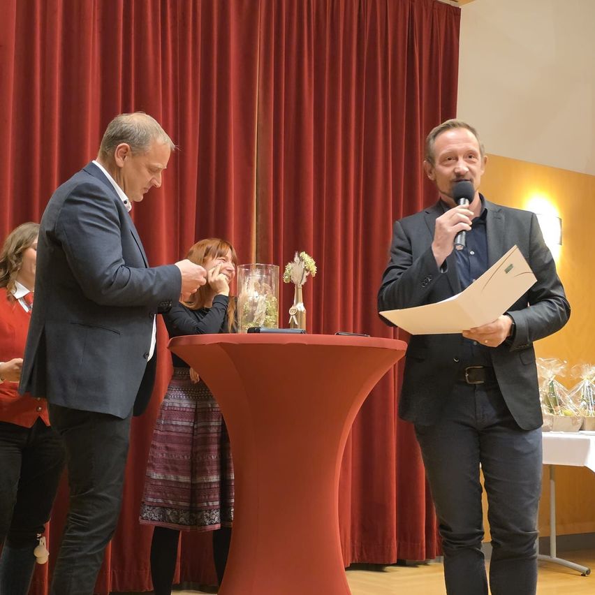 Zwei Männer stehen auf einer Bühne, einer hält ein Mikrofon und Papier, der andere eine Blume in einer Vase. Eine Frau berührt die Vase. Dahinter ein roter Vorhang und ein Tisch mit einer Blume.