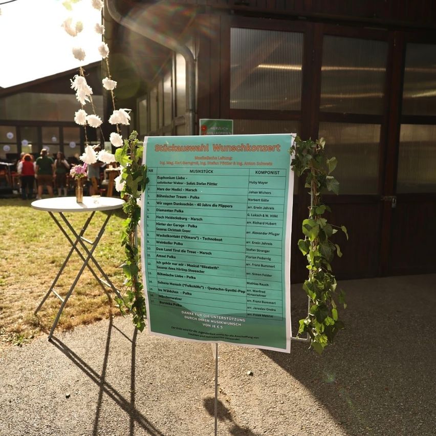 A green poster with a list of musical pieces and composers is displayed on a stand outside. The poster is decorated with green leaves and white flowers. In the background, there are people standing and sitting in front of a building with glass walls.